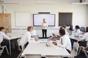 Female High School Teacher Standing Next To Interactive Whiteboard And Teaching Lesson To Pupils Wearing Uniform